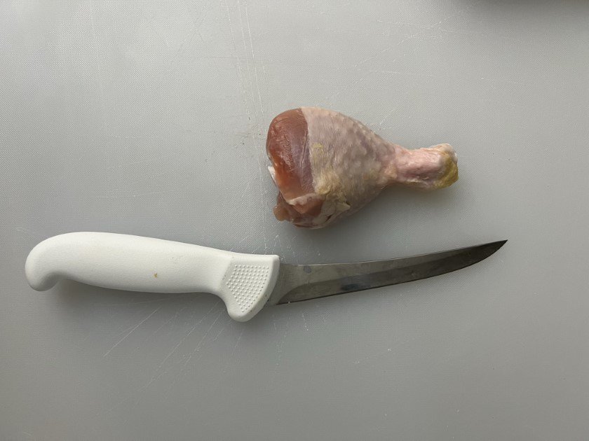 Chicken leg and boning knife laying on a white plastic cutting board.