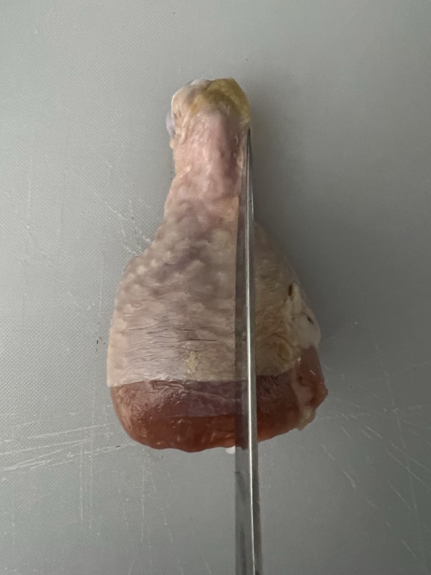Chicken drumstick about to be cut with a knife on a cutting board.